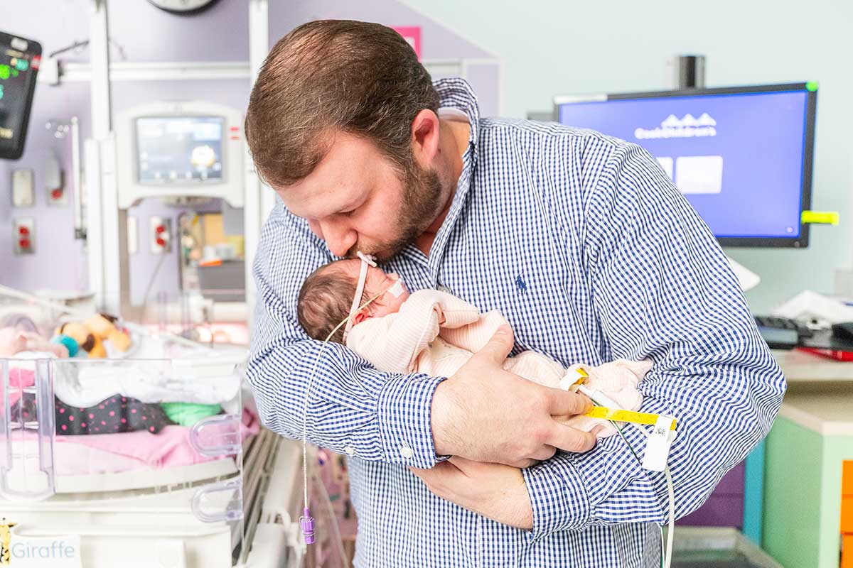 A father with a beard gently kisses his newborn baby, who has medical tubes, in a hospital's NICU