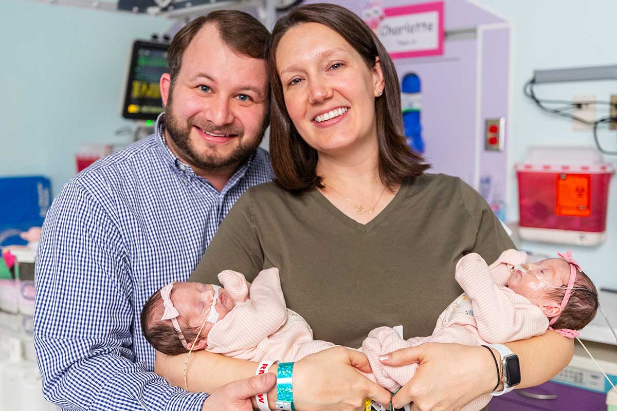 Smiling parents hold two baby girls, one with a nasal cannula, in a NICU. A sign reads Charlotte