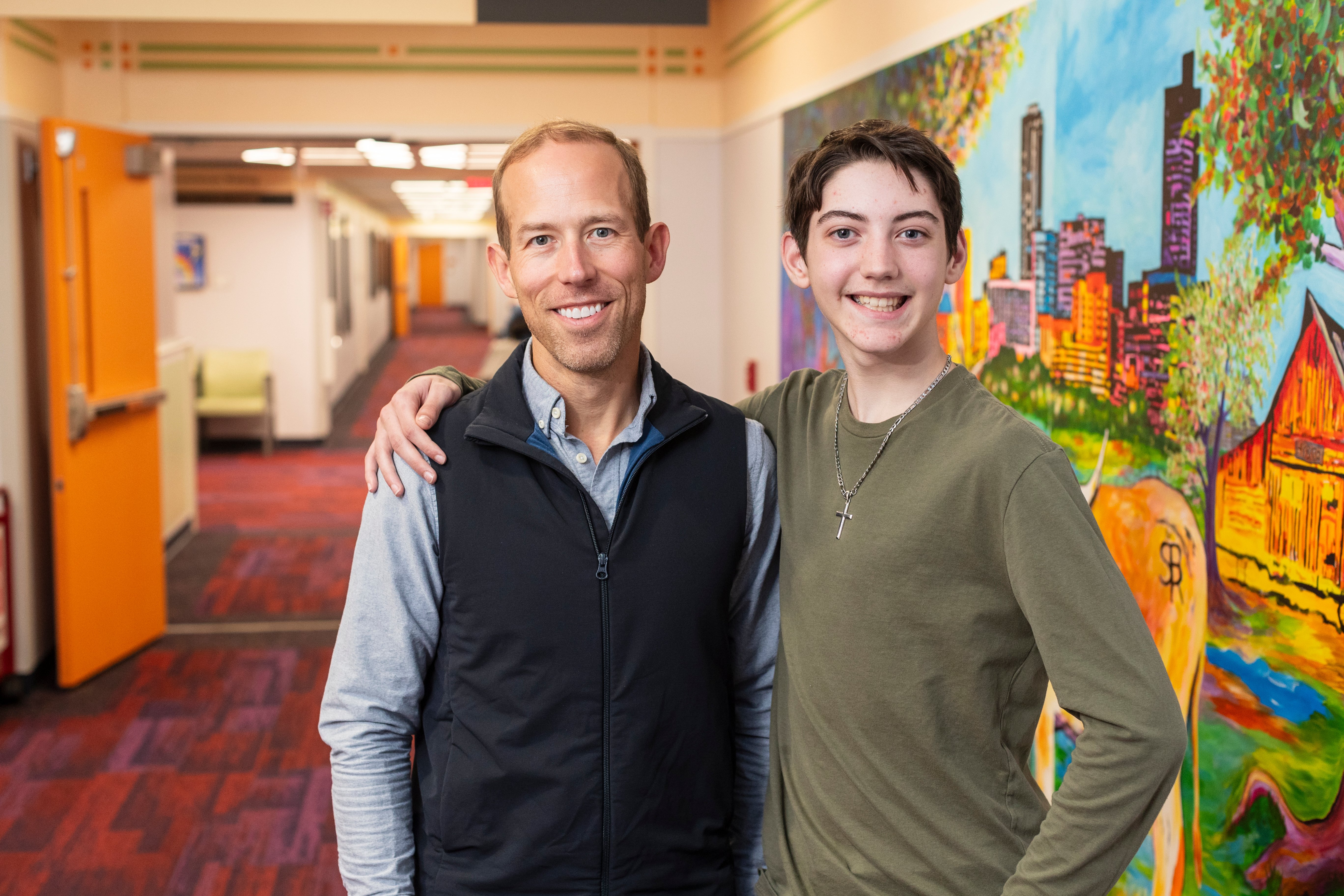 Young man wearing a green shirt has his arm around an older man wearing a blue shirt with a dark blue vest